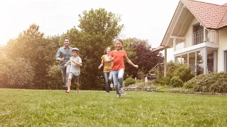 Eine glueckliche Familie mit zwei Kindern spielt im Garten vor ihrem Eigenheim.